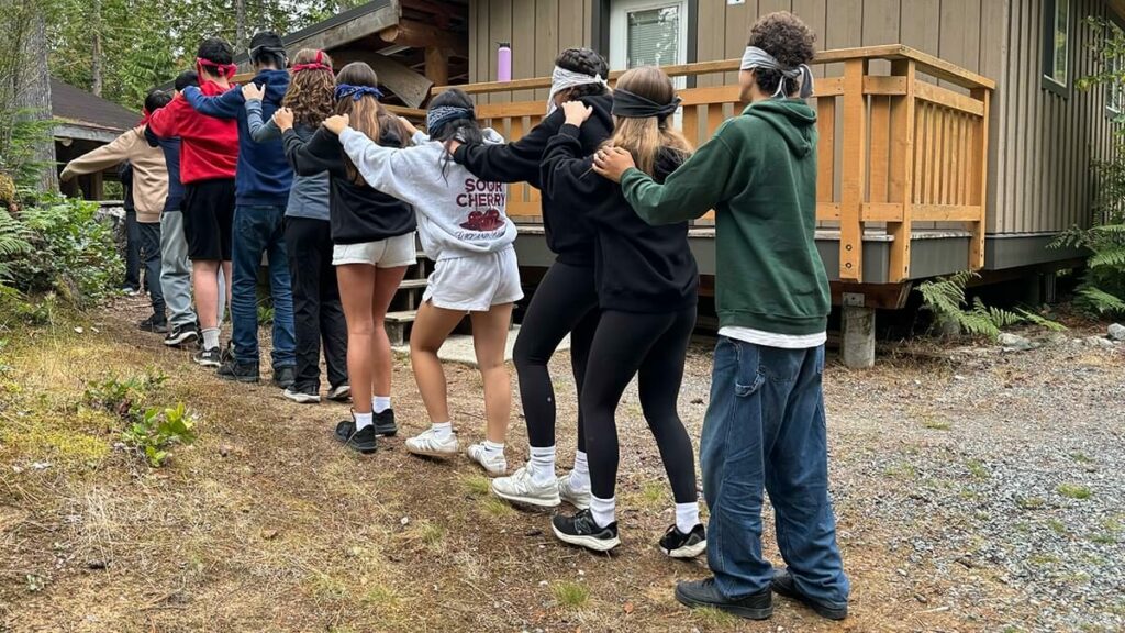 Blindfolded students line up hands on shoulders for a team-building activity outside a cabin