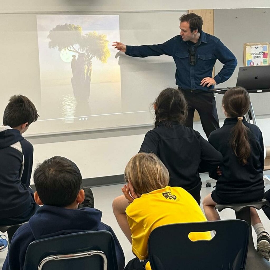 A presenter stands at the front of a classroom pointing to an image projected on the board, while a group of students sit on chairs and the floor watching attentively.