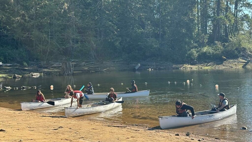 canoes with students paddle along a rocky lakeshore, all wearing life jackets