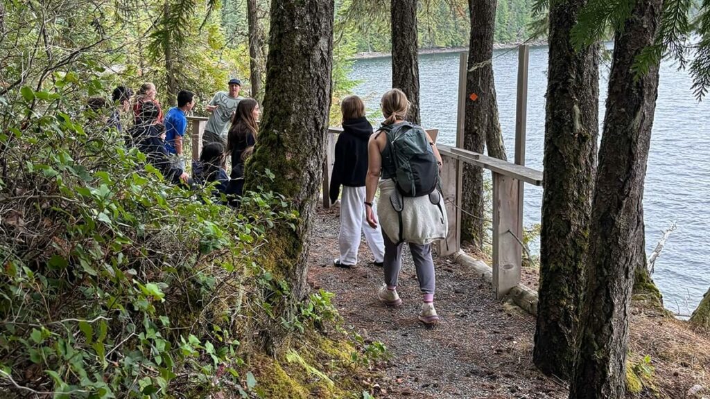 Group of students and a teacher pause on a lakeside forest trail while the teacher explains the route