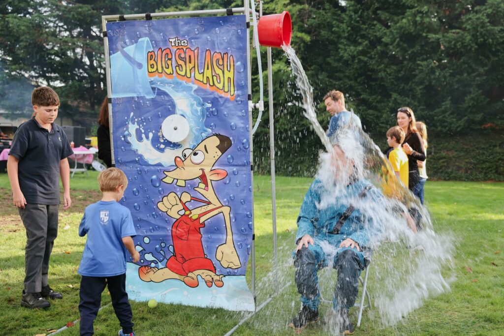 Young boy hits target at dunk tank game, pouring water over a seated volunteer during the GNS Family Carnival.